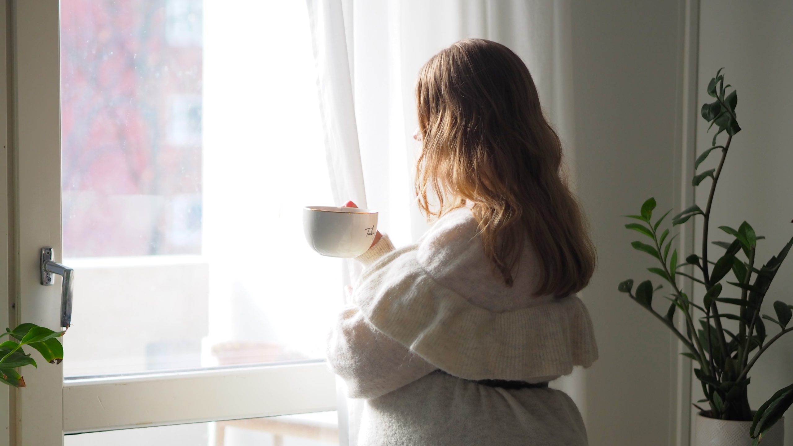 Mykkäkoulu on henkistä väkivaltaa. Oivallus Akatemia. Hanna Poikkilehto. Kuvassa villaneuleeseen pukeutunut pitkähiuksinen nainen katsoo ikkunasta ulos teekuppi kädessä.